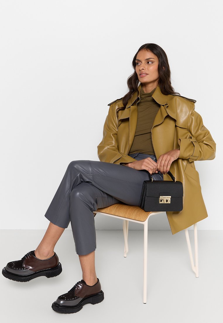 Frau sitzt auf einem Stuhl und trägt einen olivgrünen Trenchcoat, ein grünes Oberteil, graue Hose, hält eine schwarze Handtasche und trägt dunkle klobige Schuhe, während sie nach rechts schaut.