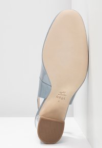 Sininen korkokenkä, jossa on sileä nahkapinta ja avoimet sivut. Vaaleanbeige ulkopohja, jossa on kuvioitu pito. Logo painettuna sisäpohjaan.