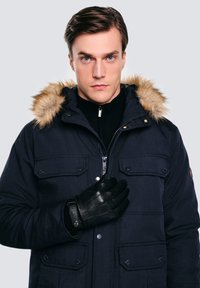 Granatowa parka z futrzanym kapturem, wyposażona w kieszenie zatrzaskowe i zamek błyskawiczny. Model ma na sobie czarne skórzane rękawiczki oraz bluzkę zapinaną na zamek pod spodem.