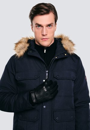 Granatowa parka z futrzanym kapturem, wyposażona w kieszenie zatrzaskowe i zamek błyskawiczny. Model ma na sobie czarne skórzane rękawiczki oraz bluzkę zapinaną na zamek pod spodem.