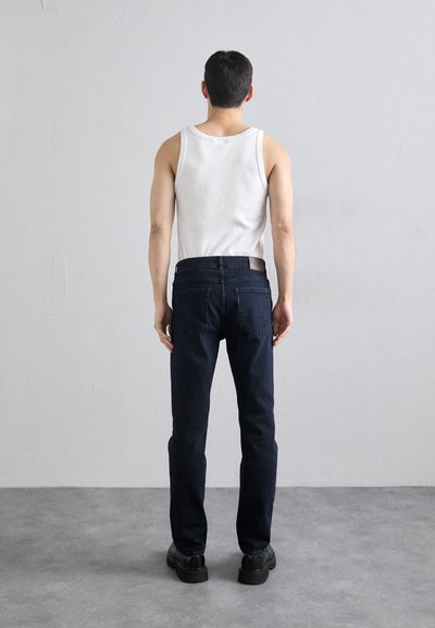 Jeans de mezclilla azul oscuro con corte recto, combinados con una camisa sin mangas blanca. Diseño simple con bolsillos traseros y pasadores para cinturón.