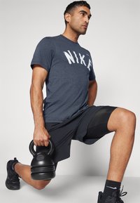 Vīriešu tumši zilas īsām piedurknēm T-krekls ar baltu "NIKE" apdruku, ap kombinēts ar melniem šortiem. Viņš tur melnu kettlebellu, veicot izklupienu.