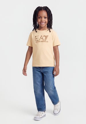 Dziewczyna z zaplecionymi włosami, uśmiechnięta, ubrana w beżowy t-shirt Emporio Armani, niebieskie dżinsy i białe trampki, stojąca z jedną nogą wysuniętą do przodu.