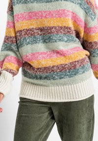 Jersey de punto con franjas multicolores en rosa, amarillo, marrón y verde azulado, con un dobladillo y puños acanalados en color crema. Combinado con pantalones de pana verdes.