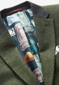 Blazer en laine verte avec un col foncé, featuring une doublure bleue à motifs affichant des designs de tickets vintage et un ruban en teal.