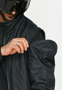 Schwarze Steppweste mit einem Reißverschluss, ausgestattet mit einer strukturierten Oberfläche und genähten Details. Eine Hand passt den Ärmel an und zeigt eine innere Schicht.