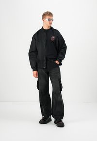 Młody mężczyzna w czarnej kurtce bombowcu, czarnym t-shircie Rolling Stones, szerokich czarnych spodniach cargo, masywnych czarnych butach i białych okularach przeciwsłonecznych, stojący.