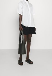 Person wearing a white short-sleeve button shirt, black shorts, black sandals, holding a black bucket-style handbag with a wide strap.