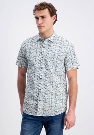 Man draagt een kortarmig overhemd met een blauw-grijs geometrisch patroon, gecombineerd met donkerblauwe spijkerbroek, staand tegen een lichte achtergrond.