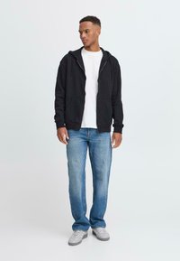 Svart hettegenser med glidelås og frontlommer, brukt over en hvit t-skjorte, kombinert med lyseblå jeans og grå joggesko, stående mot en enkel bakgrunn.