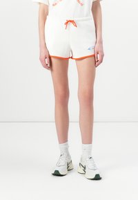 Pantaloni corti bianchi con bordino arancione e vita con cordino, con un piccolo logo blu sul lato, indossati con calze bianche e scarpe da ginnastica.