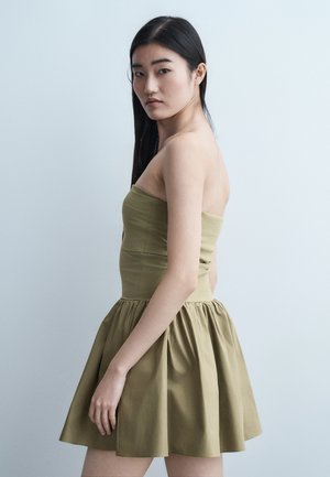 Jauna moteris su ilgais tamsiais plaukais, vilkinti alyvuogių žalios spalvos be petnešėlių suknelę, stovinti šonu priešais paprastą šviesų foną.