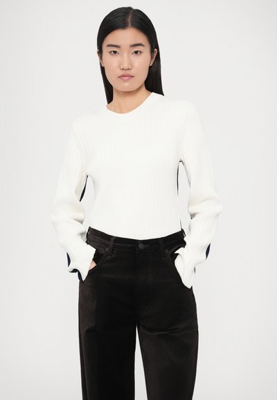 Bluză albă cu mâneci lungi, cu model tip rețea și gât rotund, asortată cu pantaloni negri din catifea de bumbac, cu talie înaltă. Mâinile sunt în buzunare, fundal neutru.