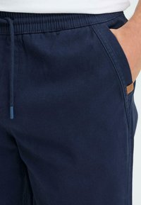 Un pantalon de jogging bleu marine avec une taille élastique munie d'un cordon de serrage, des poches latérales et une petite étiquette logo marron sur la zone de la poche.