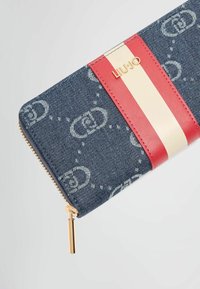 Portafoglio in denim con hardware dorato, accenti a strisce rosse e crema e un motivo logo ripetuto. Presenta una chiusura a zip e un contrasto testurale.