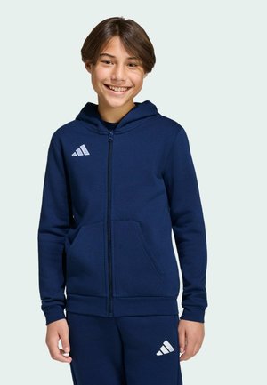 Uśmiechający się młody chłopiec ubrany w granatową bluzę z kapturem i suwakiem oraz pasujące spodnie z białymi logo marki sportowej.