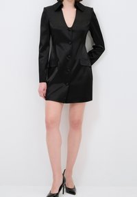 Femme portant une robe blazer en satin noir ajustée avec des boutons sur le devant et des escarpins noirs, debout devant un fond blanc.