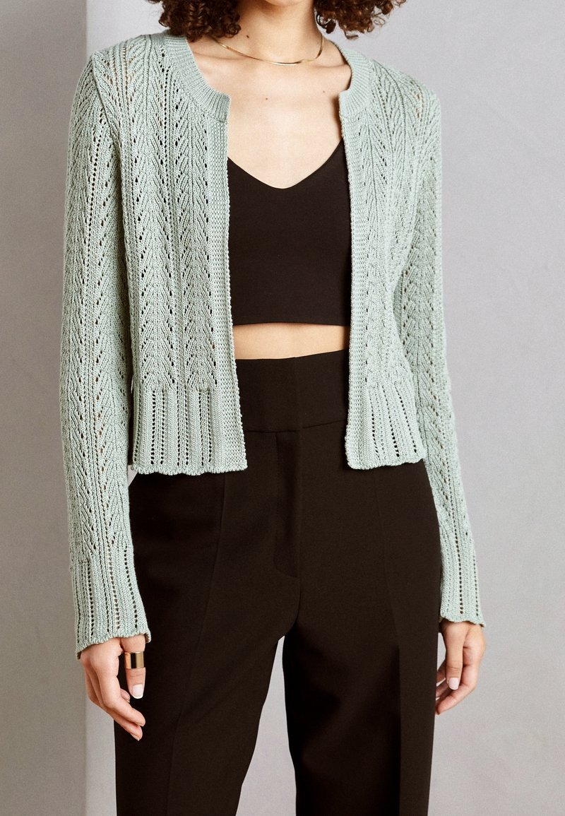 Femme portant un cardigan tricoté vert clair sur un top court noir et un pantalon noir taille haute, debout devant un fond uni.