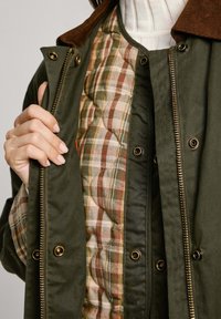 Veste vert olive avec un col en velours côtelé marron, zips dorés et doublure matelassée à carreaux dans des tons terreux. Comprend des boutons-pression et un tissu texturé.