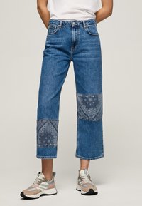 Jeans i denim med ljusblå färg, bred benstil och mönstrade blå lappar på knäna. I kombination med flerfärgade sportskor.