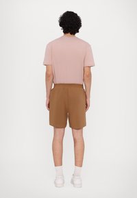 Brune shorts med elastisk midjebånd og rosa t-skjorte, kort erm. Hvite joggesko og ankelstrømper synlige, sett fra baksiden.