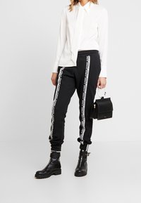 Černé joggingové kalhoty s bílými bočními pruhy, v kombinaci s bílou košilí s kravatou, černými kotníkovými botami a malou černou kabelkou.