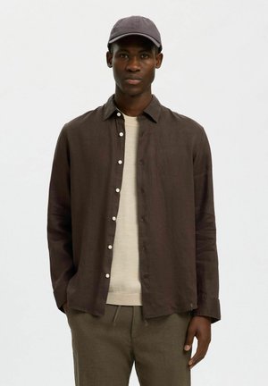 Jeune homme portant une chemise marron foncé boutonnée par-dessus un pull beige, un pantalon olive et une casquette foncée, debout devant un fond uni.