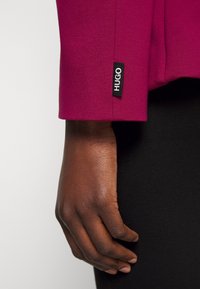 Main à la peau foncée suspendue à côté d'une personne portant un manches longues magenta avec une étiquette de marque noire "HUGO" près du poignet et un pantalon noir.