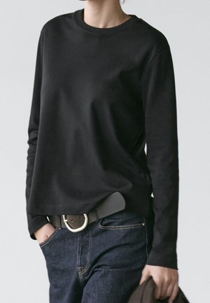 Osoba ubrana w prostą czarną koszulę z długim rękawem, ciemnoniebieskie dżinsy z czarnym paskiem, trzymająca w jednej ręce ciemną kurtkę.