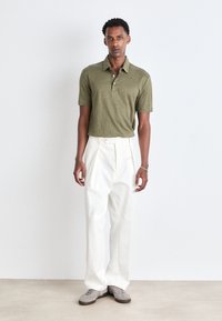 Olivgrön polo med krage, tillsammans med vita plisserade byxor. Modellen bär grå mockaskor och står framför en neutral bakgrund.