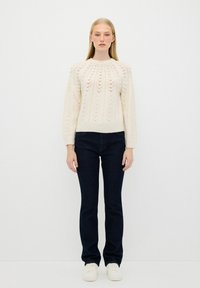 Maglione lavorato a maglia crema con un motivo testurizzato, scollo rotondo e maniche lunghe, abbinato a jeans blu scuro e sneakers bianche.