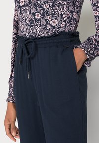 Granatowe spodnie o luźnym kroju z elastycznym pasem i sznurkiem, z bocznymi kieszeniami i gładką fakturą. Zestawione z bluzką w kwiatowy wzór.
