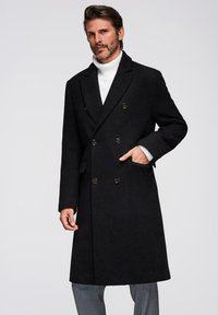Abrigo negro de lana de doble botonadura con botones de latón, bolsillos laterales y cuello con muesca, superpuesto a un jersey de cuello alto blanco.