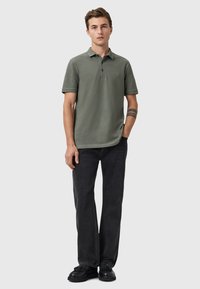 Olivgrünes Polohemd mit kurzen Ärmeln, Kragen und kleinem Logo auf der Brust; kombiniert mit dunklen Jeans und schwarzen Schuhen. Einfaches, tailliertes Design.