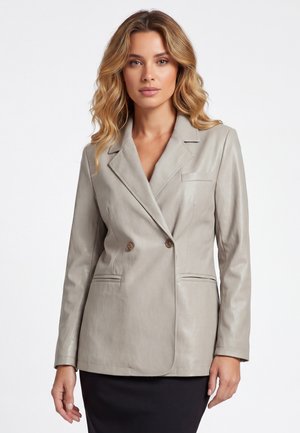 Vrouw met golvend blond haar die een lichtgrijze dubbelgeknoopte leren blazer en een zwarte rok draagt, staand tegen een witte achtergrond.