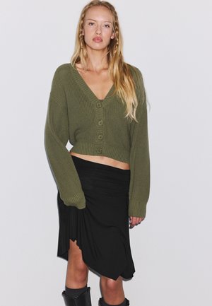 Femme blonde portant un cardigan court boutonné vert olive, une jupe asymétrique noire et des bottes noires sur un fond clair uni.