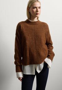 Pull en maille marron avec un ourlet côtelé et des manches larges, superposé sur une chemise blanche à boutons, porté avec un jean en denim foncé.
