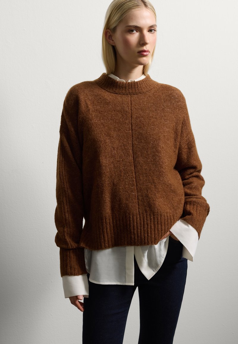 Pull en maille marron avec un ourlet côtelé et des manches larges, superposé sur une chemise blanche à boutons, porté avec un jean en denim foncé.