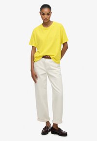 Gul kortærmet t-shirt parret med opkrøllede cremede bukser og brunt vævet bælte. Modellen har mørke loafers på og står foran en neutral baggrund.