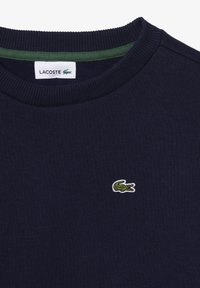 Tmavomodrý bavlnený sveter s rebrovaným okrúhlym výstrihom. Obsahuje malé zelené a biele logo krokodíla Lacoste na hrudi.