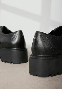 Chaussures en cuir noir avec un relief texturé et une semelle épaisse à relief. Présentent des accents métalliques circulaires et un décor brogue le long des bords.