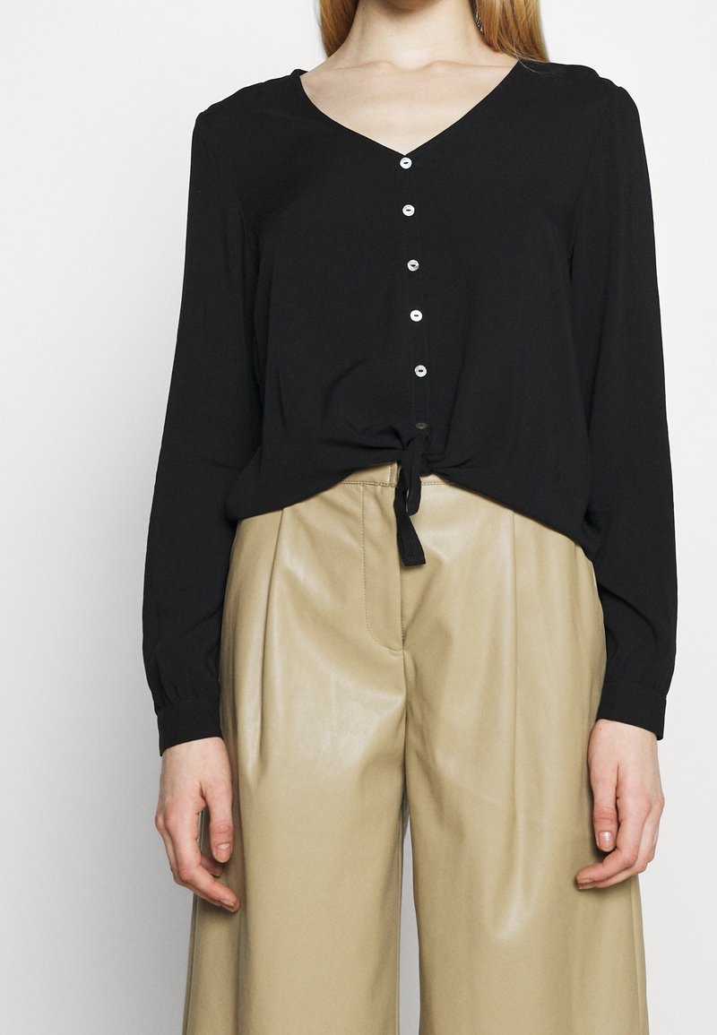 Blusa nera a maniche lunghe con scollo a V, chiusura con bottoni e nodo in vita. Abbinata a pantaloni beige in ecopelle a vita alta.