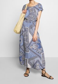 Robe longue en tissu bleu à motifs, manches courtes, taille élastique et jupe fluide. Associée à des sandales plates noires et un sac fourre-tout beige.