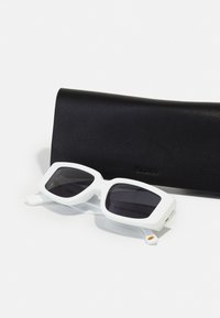 Gafas de sol blancas con un armazón grueso y angular y lentes oscuros, situadas junto a un estuche de cuero negro con un acabado texturizado.