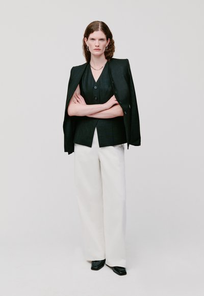 Schwarze taillierte Jacke mit strukturierten Schultern über einem dunklen, geknöpften Weste; kombiniert mit lockeren weißen Hosen und schwarzen viereckigen Schuhen.