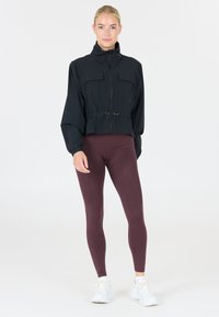 Black cropped jacket with a high collar, two front pockets, cinched waist, paired with maroon leggings and white sneakers. Smooth texture.