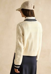 Kreemikootud kardigan, millel on tekstureeritud muster, soonikserv ja tumesinised triibulised käised. Kombineeritud tumeda seeliku ja beeži korkiga.