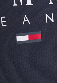 Tissu côtelé bleu marine avec des lettres blanches et un logo rectangulaire en blanc, rouge et bleu marine. Probablement une partie d’un vêtement de marque.