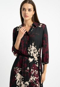 Kobieta z długimi brązowymi włosami, ubrana w czarną i bordową sukienkę w kwiaty z kołnierzykiem i czarnym paskiem w talii, spoglądająca w lewo.