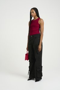Red ribbed sleeveless top, black cargo pants with zippers, and pointed black heels. Holding a small pink handbag. Simple background.